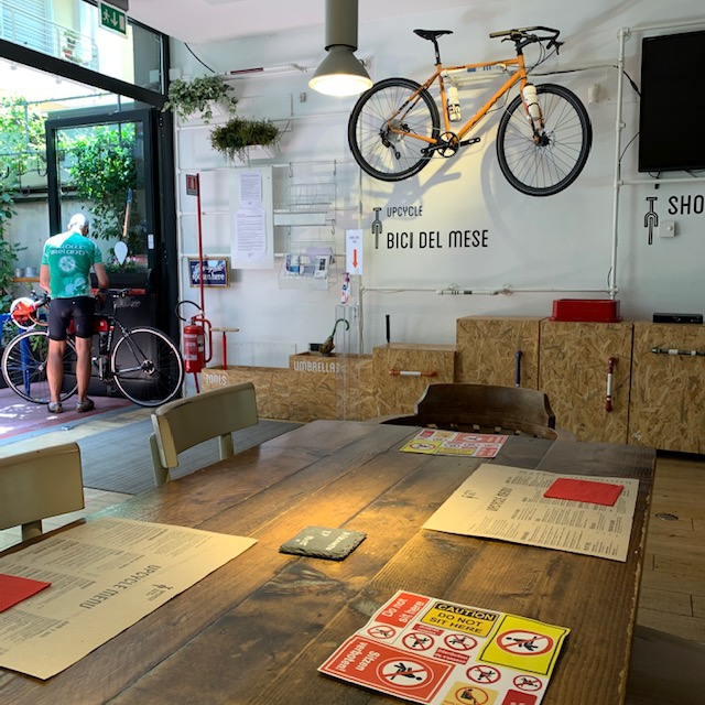 最後にご紹介するのは、自転車カフェ、Upcycle cafe Milano。週一で無料自転車修理のサービスを行なっており、また修理の仕方も見せてくれるので、学んで自分で修理することも可能になるとのことだ。自転車で旅をした人が自分の経験を語るイベントや自転車に関する本の出版イベントなどなども定期的に開催されて自転車好きが集まるスペースとなっている。
カフェレストランの奥にはシェアオフィスのスペースもある。コロナの影響で利用客は減ったそうだが、カフェで飲み物を飲みながら仕事をしたり勉強している人もいた。ここでもソーシャルディスタンスが守られていて、まずカフェに入る前に体温チェック。座る席は写真のように隣や向かいに座れないようになっている。カフェのメニューにはイタリア語ではない外国語表記が。「この会社で働く人達は多国籍で、シェフがスウェーデン人なのでスウェーデン料理を中心に提供しているんだよ。自転車が人を繋ぐカフェだけど、国際性を出すことで他の場所と差をつけたくてね。」とフランス人のフロアマネージャー、マチュー君が教えてくれた。朝早くから夜遅くまで営業していて、気が向いた時にいつでも立ち寄ることができる場所だ。自転車で移動するミラノ。歩いたり公共交通機関よりも早く移動できて、運動にもなって、今後ミラネーゼ達のライフスタイルがより健康的で環境に配慮したものになる予感がしている。