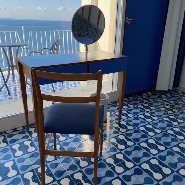 部屋の鏡付きテーブル、ヴァニティにはもちろんジオ・ポンティの代表作である椅子スーペルレッジェーラ。全てがブルー。そして部屋の窓からは空と海のブルーが果てしなく広がる。
「ソレントのホテルを作ったとき、必要性はなかったのだが、100室ある客室をそれぞれ違う床にしたいと思った。これは、わたしの陶器への愛情からくるもので、それが生かされると分かると頼まれたこと以上のことをしなければと背中を押されるのだ。」とはジオ・ポンティの言葉。33種類のタイルをデザインしたが、そのうち5つは作られず、5種類はホールやレストランなどの公共スペースに使用され、残りの23種類が客室に使われている。タイルの模様に合わせてそれぞれの部屋の壁画とクッションの模様も異なる。彼のこだわりある斬新なデザインは、時代を越えて今でも充分新しく実にオシャレだ。